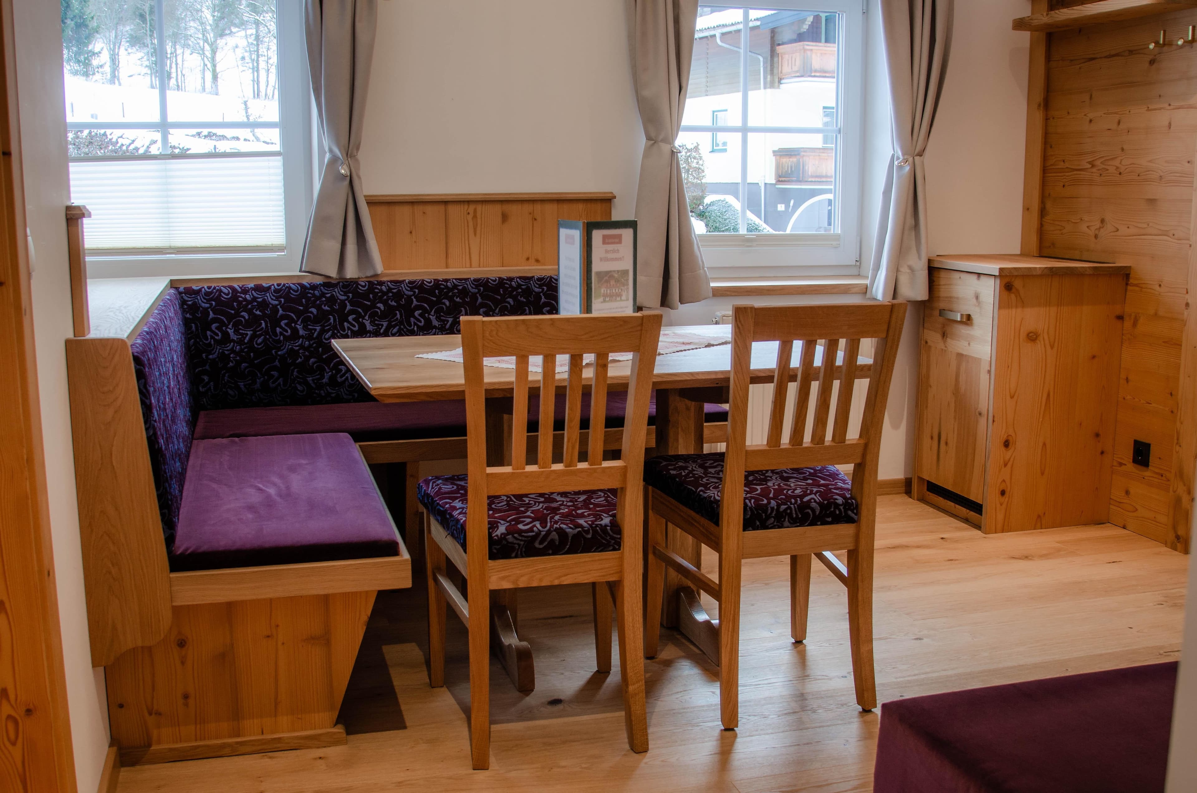 Bauernhof Krahlehen · Familiensuite mit Frühstück - A bedroom featuring a double bed with a light wooden frame and a dark purple patterned headboard. The bed is neatly made with crisp white bedding and prominent large white pillows. A small wooden nightstand with an open shelf and an adjustable reading lamp are visible on one side. The room is brightly lit, offering a clean and inviting look.