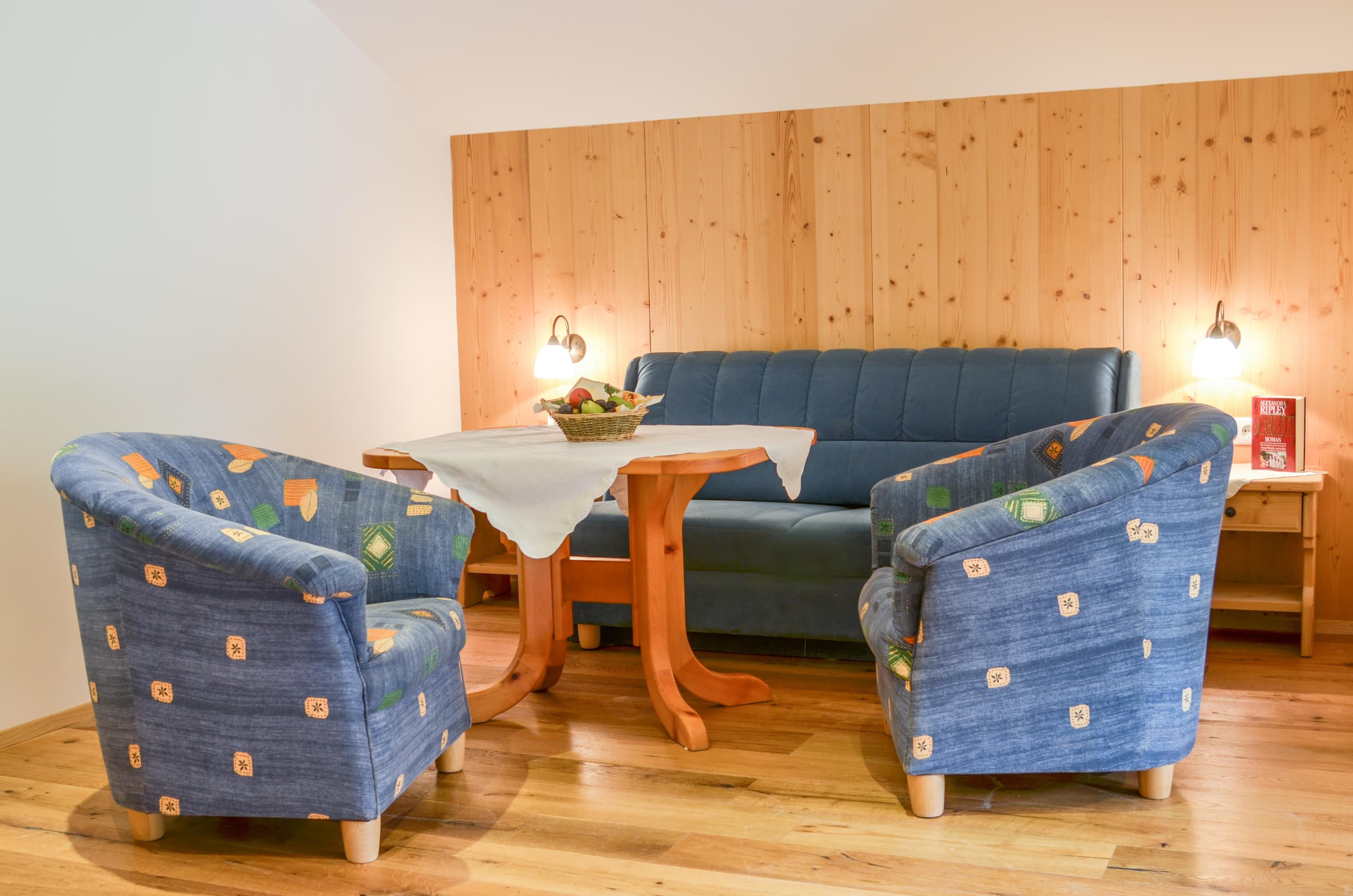 Bauernhof Krahlehen · Apartment Top 1 - A bright and peaceful bedroom featuring a comfortable wooden double bed with two single mattresses, flanked by wooden side tables with reading lamps. A wooden bench sits at the foot of the bed. A window with floral curtains allows natural light to illuminate the wooden floor and white walls, creating a serene atmosphere.